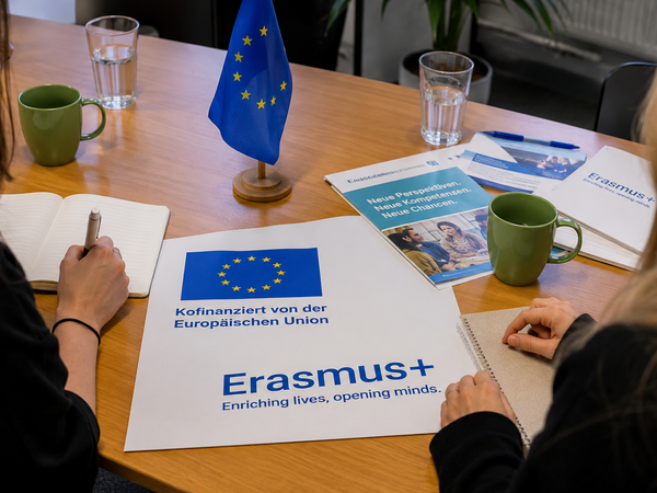 Frauen sitzen an einem Tisch und in der Mitte liegt ein Plakat mit Erasmus + und Kofinanziert von der Europäischen Union