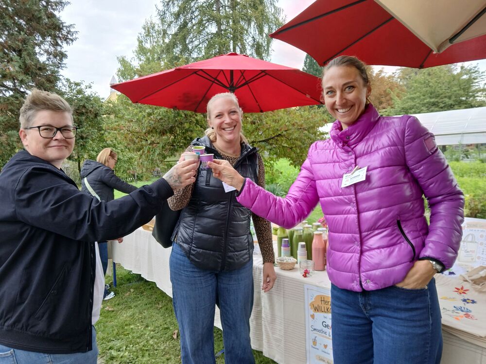 Drei Kolleginnen stoßen mit Smoothie-Bechern an und lächeln in die Kamera
