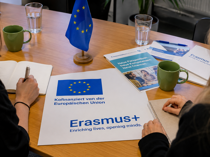 Frauen sitzen an einem Tisch und in der Mitte liegt ein Plakat mit Erasmus + und Kofinanziert von der Europäischen Union